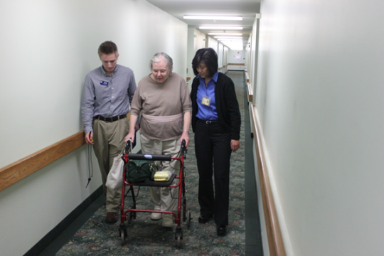 Physical therapy student and professor lead senior community resident down hallway