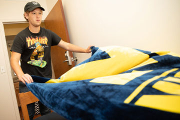 A student is placing a blanket on his bed in his dorm room.