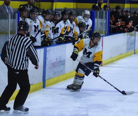 A UM-Flint Men's Club Hockey player handles the puck while a referee skates nearby and teammates watch from the bench.