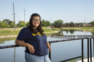 UM-Flint Student Government President Samantha Uptmor poses for a portrait on a bridge over the Flint River. She is wearing a blue UM-Flint T-shirt.