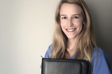 Quinn Hanses poses for a portrait. She is wearing blue nursing scrubs and is holding a UM-Flint School of Management padfolio.