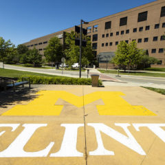 The UM-Flint logo (Block M in yellow with Flint underneath in white) on the sidewalk in front of a building on campus.