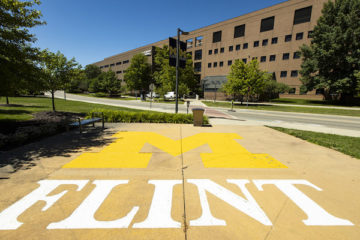 The UM-Flint logo (Block M in yellow with Flint underneath in white) on the sidewalk in front of a building on campus.