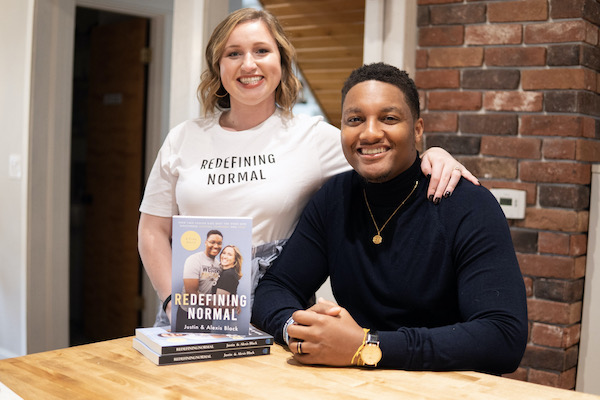 Alexis Lenderman-Black and her husband, Justin Black, pose for a portrait. They sit at a table with a copy of their book, Redefining Normal.