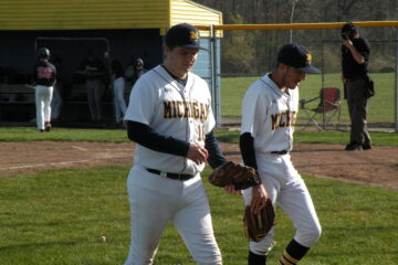 Two male UM-Flint students wearing white baseball uniforms with blue hats and carrying baseball gloves walk out onto a baseball field with a dugout in the background.