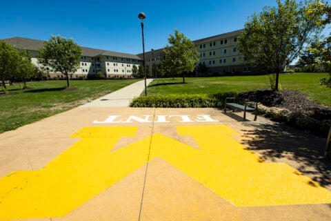 UM-Flint's First Street Residence Hall in the background on a summer day with the Block M logo prominently in front on the sidewalk.