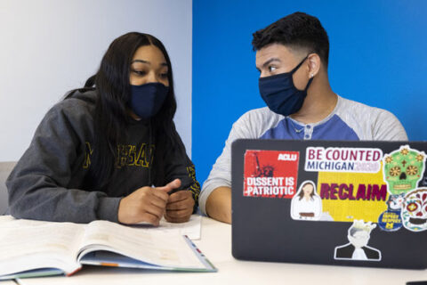 Two UM-Flint students, one woman and one man, sit and talk at a table with a laptop and notebook.