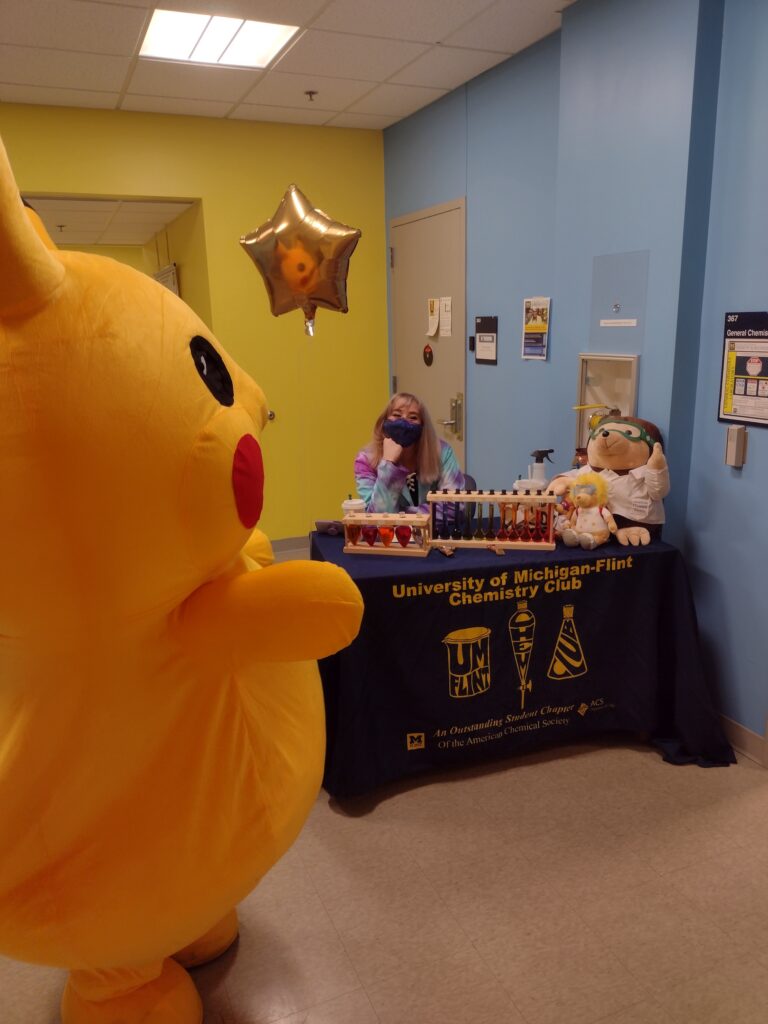 Student in large Pikachu costume poses with Chemistry Club informational table 