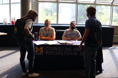 Two UM-Flint students talk to CAPS Social Workers Kayla Bueby and Jenna Newton, who are seated at a table in the UCEN.