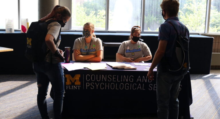Two UM-Flint students talk to CAPS Social Workers Kayla Bueby and Jenna Newton, who are seated at a table in the UCEN.