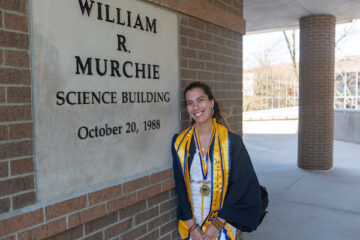 Nicole-Kristine Smith, in a graduation robe and regalia, poses in front of the William R Murchie Science Building sign