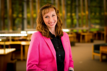 Tischler stands for a waist-up photo with bangs, shoulder-length light-brown hair, a big, kind smile, a black shirt, and pink blazer. There are desks in the distance.