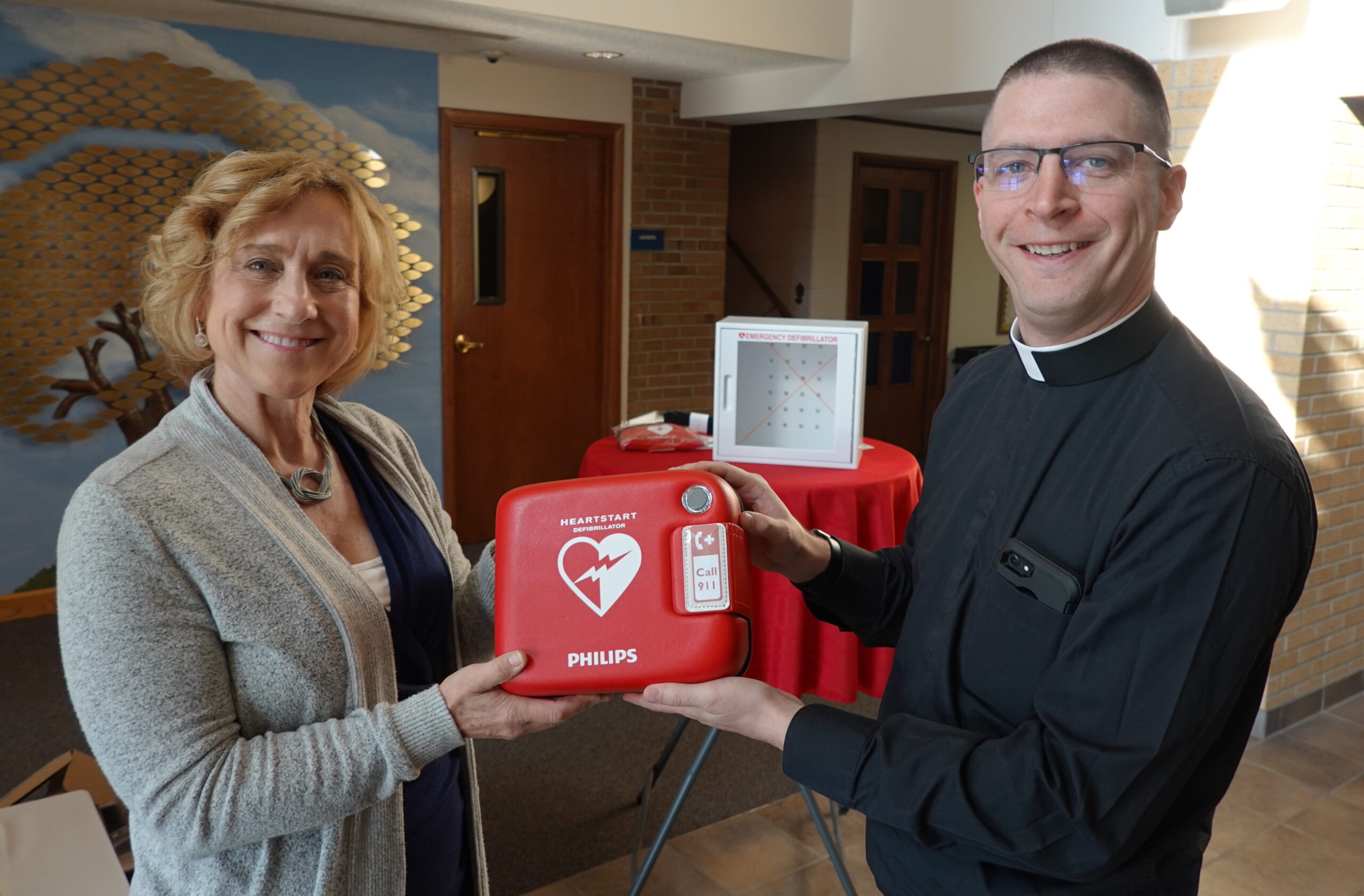 Kristi Wilson, associate professor at UM-Flint's School of Nursing presents Father Anthony of Pius X Catholic church a new, automatic defibrillator.