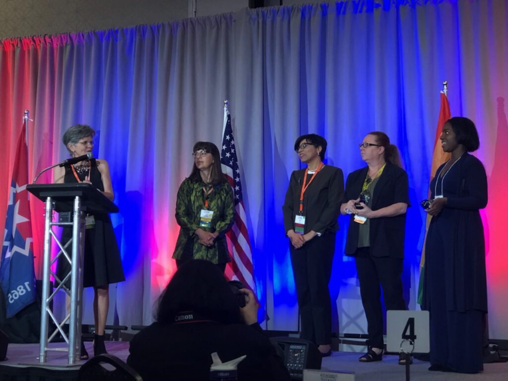 Silva (second from right) and Wilson (far right) receiving the award at the 2022 MI-ACE conference. Mary Jo Sekelsky, executive director of alumni relations at UM-Flint, is at the podium