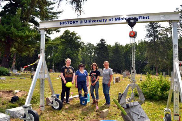 The UM-Flint Cemetery Preservation team (pictured right to left: Lisa Horn of Novi, Wendy Kimberly of Fort Gratiot, Izzy Guertin of Fenton and Mary Ann Kost of Linden) pose in front of a Gantry crane at Old Calvary Catholic Cemetery in Flint