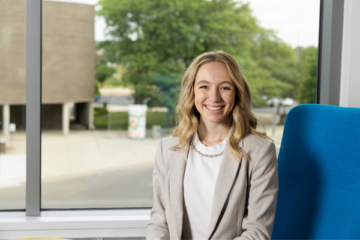 Culinski poses on campus for a professional headshot. She is seated, in a sharp suit, and has shoulder length blonde hair surrounding a genuine smile.