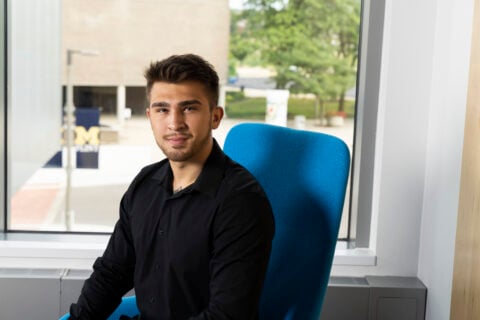 Nathaniel Cordova poses in a blue chair with the UM-Flint Rec center behind him out the window. He has a black button up shirt on and is dapper in appearance.