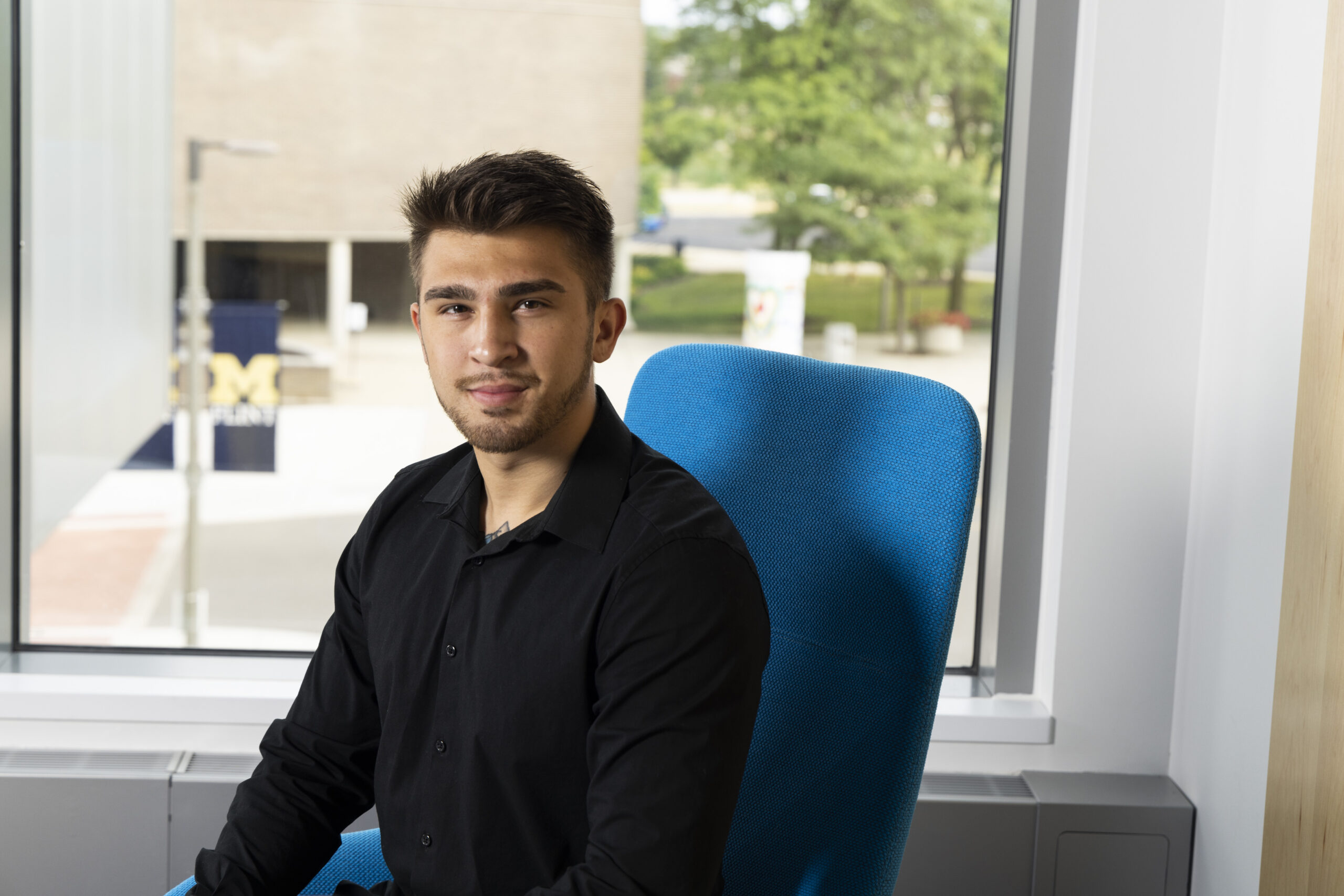 Nathaniel Cordova poses in a blue chair with the UM-Flint Rec center behind him out the window. He has a black button up shirt on and is dapper in appearance.