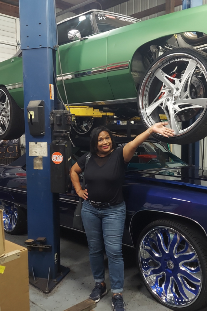 Professor Shirl Donaldson poses before a hoisted green donk and a blue convertible donk parked beneath.