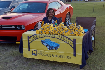 Shirl Donaldson sits behind a UM-Flint College of Innovation & Technology table loaded with T-shirts with a red Dodge Challenger GT behind her.