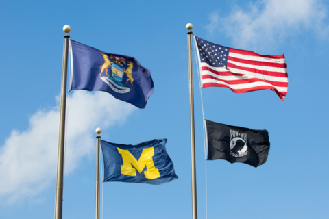 A photo of flags against a blue sky: The Michigan state flag, the University of Michigan flag, the American flag, and the POW MIA flag