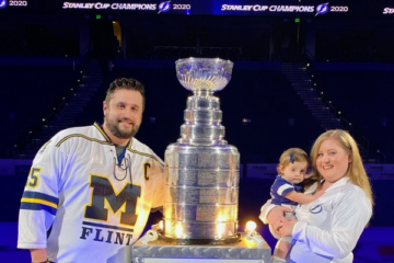 Tom Garavaglia posing with wife and baby next to the Stanley Cup in UM-Flint hockey gear