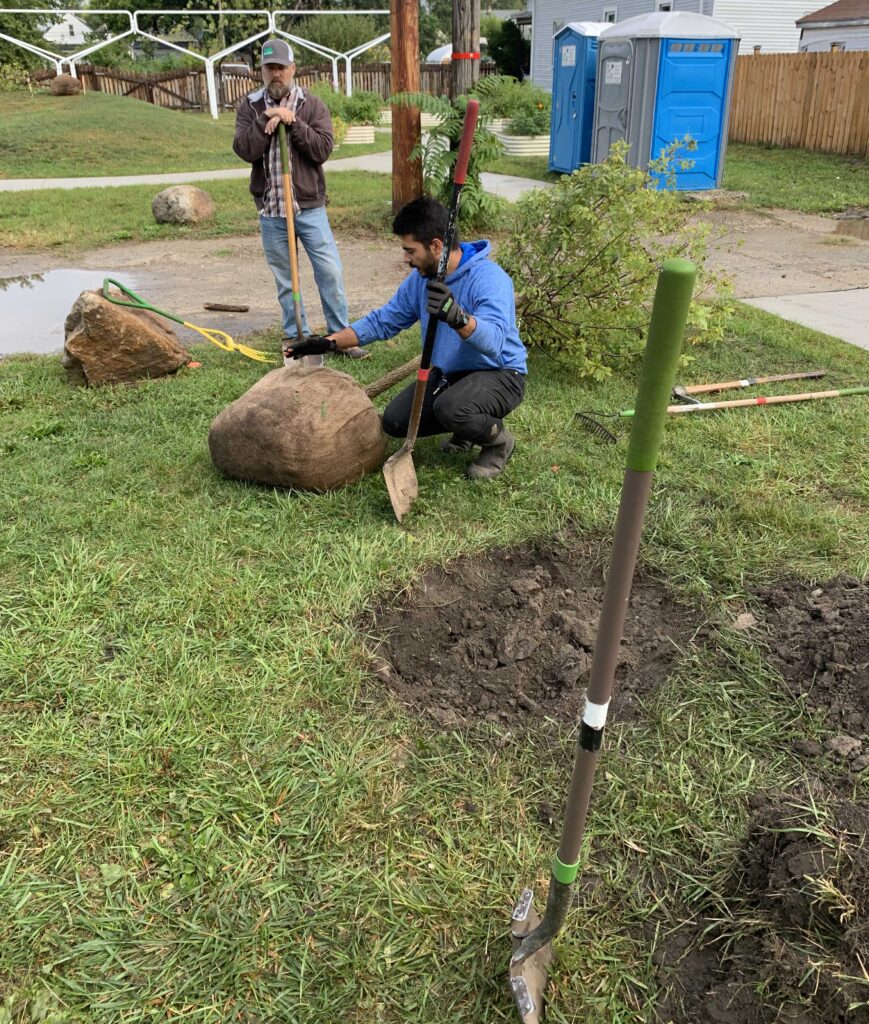 Two demonstrators show how we measure the root ball of a tree so that we dig the hole properly for the tree. One volunteer is crouched by the burlap-wrapped roots, and another stands nearby with shovel in hand.