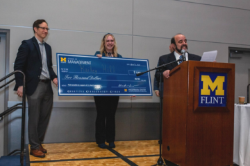 A student accepting a big check award on stage with two professors, one behind a UM-Flint podium