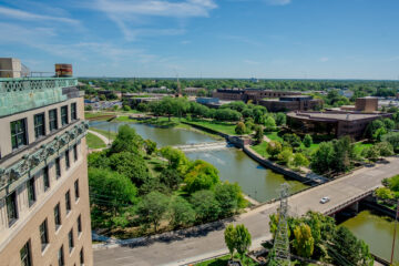 A view of UM-Flint from the Northbank Center. Thompson Library and the University Center are visible across the Flint River in summertime.