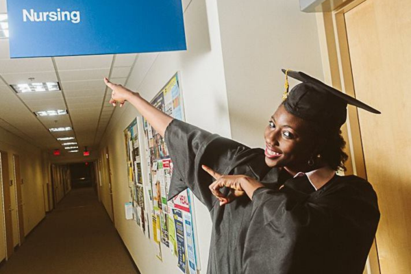 Japari Gadzama wears graduation cap and gown as she smiles and points to signage outside the nursing offices UM-Flint