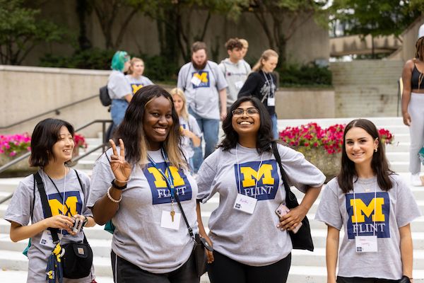 Four UM-Flint students wearing UM-Flint T-shirts smile in McKinnon Plaza on campus.