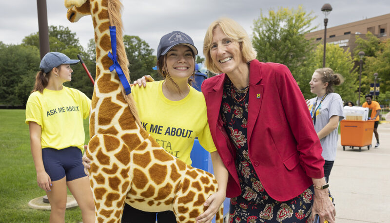 A student holding an oversized stuffed giraffe posing with Interim Chancellor Donna Fry