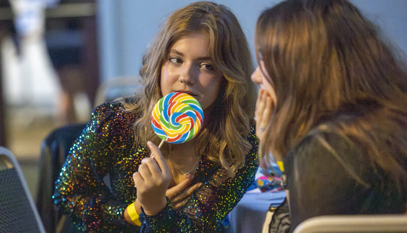 A student holding a lollipop talking to another student at the homecoming dance