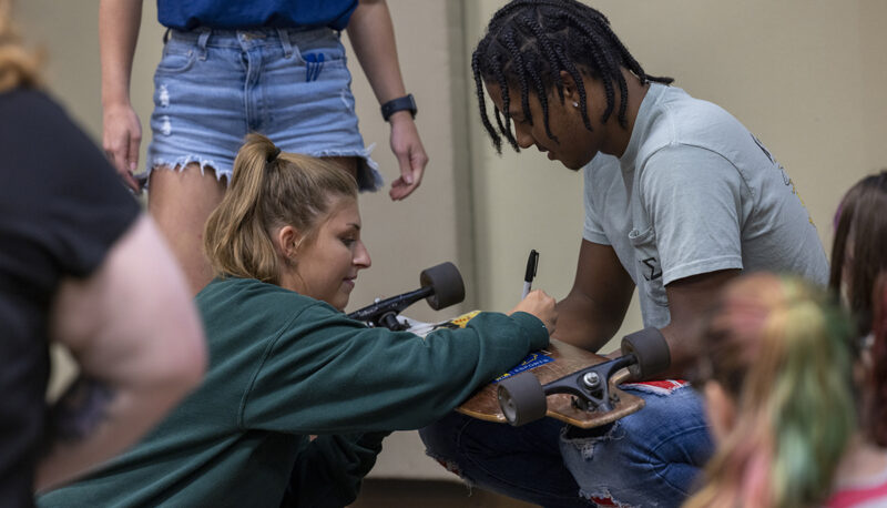 One student holding a skateboard while another signs it