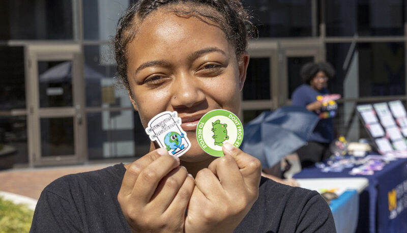 A student holding up two stickers at Eco Extravaganza: One says "Don't frog-it to recycle with a picture of a frog, the other says "Every Day is Earth Day" with a cartoon Earth mascot