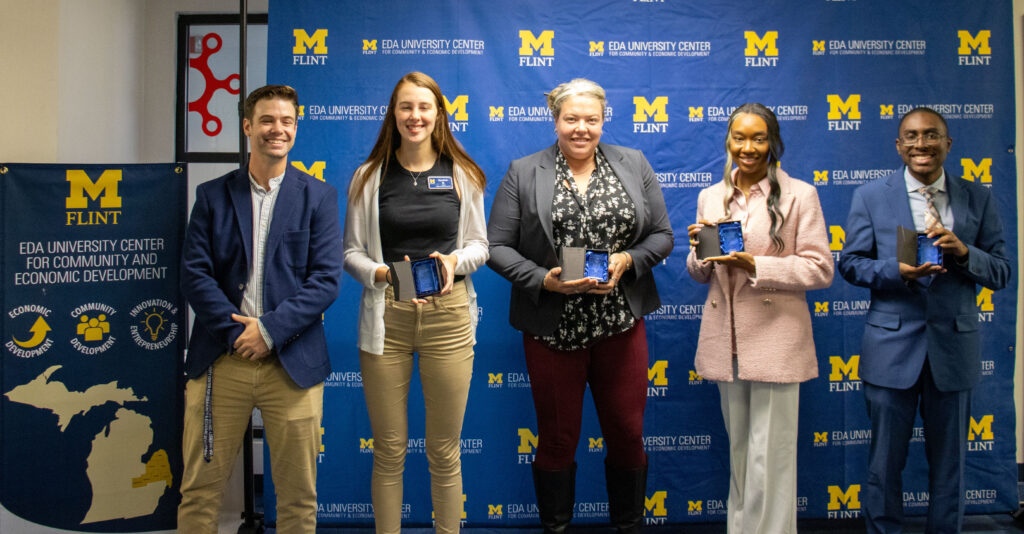 EDA University Center for Community and Economic Development sign is to the left. The presenter, and then four award recipients all stand in a line with a UM-Flint backdrop. The recipients are holding cases open with crystal awards.