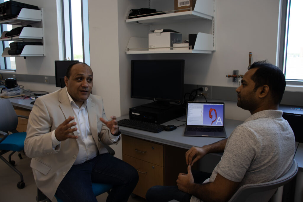 A male professor sits in a rolling chair talking animatedly with his hands to another man, also seated. Between them is a laptop with menus and writing to the left and an image to the right with a red tube in the shape of a question mark and a light blue background.