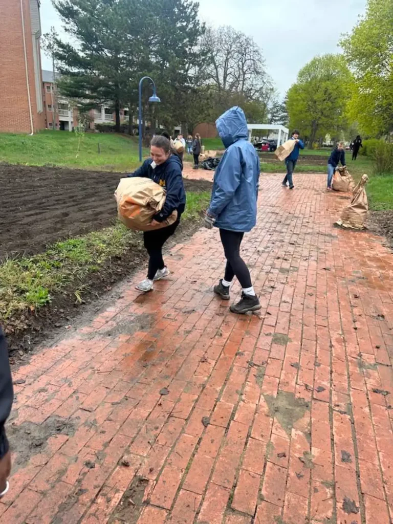 Image shows two students in the foreground laughing and carrying a refuse bag. They are standing on a brick path. To their left is a tilled garden, and there are around 6 other students and an apartment complex in the rear of the image. It appears there is a light sprinkle.