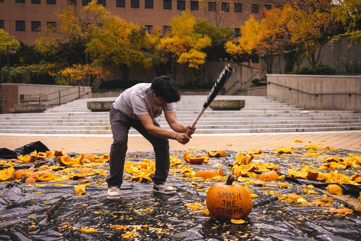 A student smashing a pumpkin with a baseball bat in McKinnon plaza. The pumpkin has the words "smash the stigma written on it."