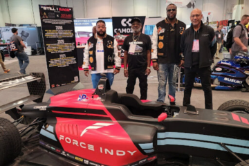 There is an IndyCar in the foreground that says "Force Indy" on the side. A tall poster is on the left behind that with the tittle "Still I race" and four men are standing behind the car posed for a picture. The rear of the image shows a setting of an automotive show at an indoor conference center.