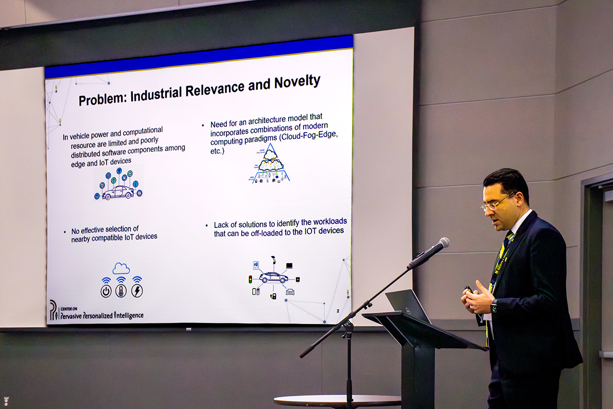 Kessentini stands to the right of the image facing a podium. Behind him, there is a large screen that says, "Problem: Industrial Relevance and Novelty" and says what some of the issues with computing in automobiles are currently, with images. Of a car, of a pyramid chart, of IoT device icons, and another showing how those devices are all part of the car's processing workload.