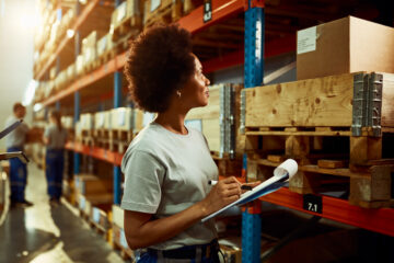 A woman with a clipboard looking at pallets in a warehouse.