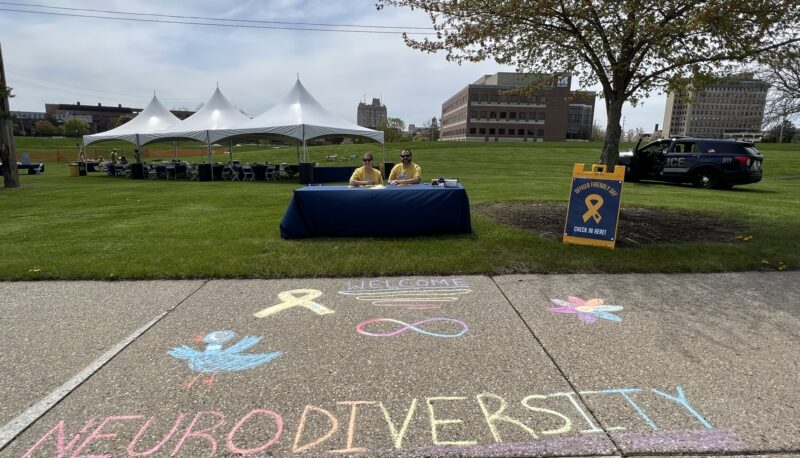 A table with two staffers behind it, and the word "neurodiversity" chalked on the sidewalk.