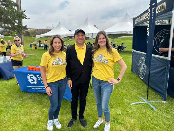 Two women pose with a man in a grassy field, marquee tents and other displays are featured behind them.