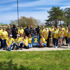 A group of people including volunteers and staff wearing yellow shirts and law enforcement officers pose together on a sunny day on a grassy field.