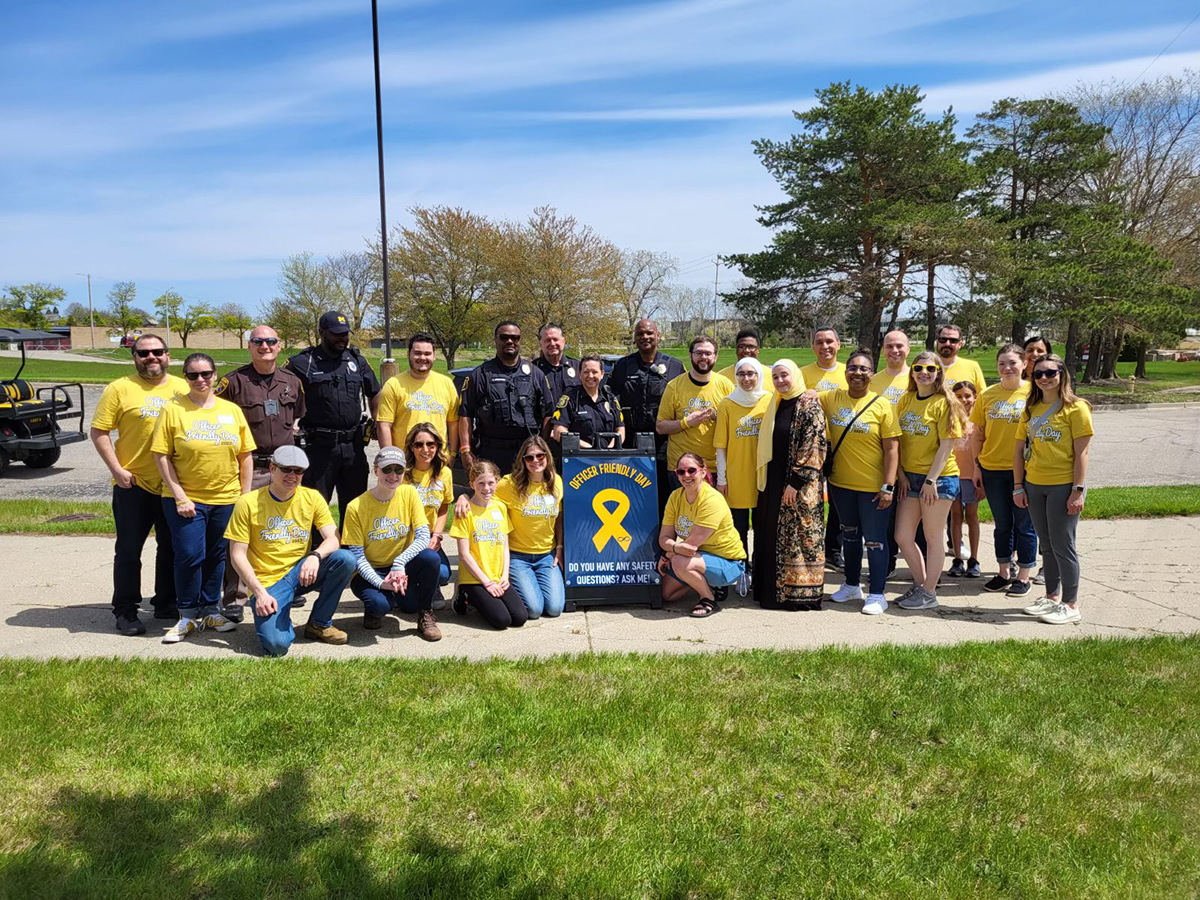 A group of people including volunteers and staff wearing yellow shirts and law enforcement officers pose together on a sunny day on a grassy field.