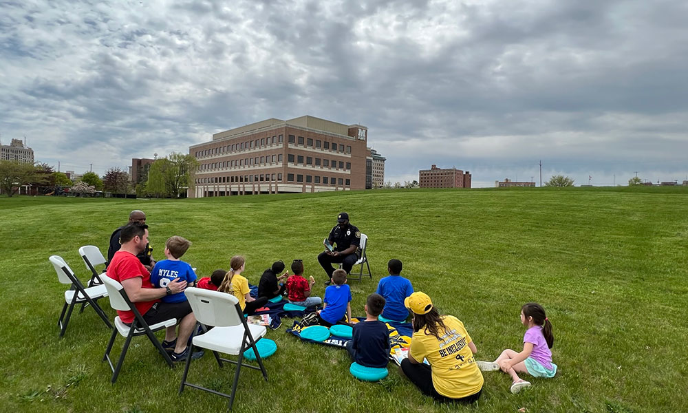 Parents and children sitting in a grassy field while a police officer reads to them. A large building is in the background with a cloudy sky.