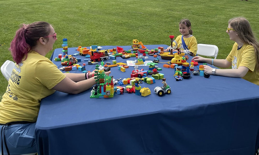 Two female volunteers play and a young girl play with toy blocks on a table with a blue tablecloth, located outside in a grassy field.