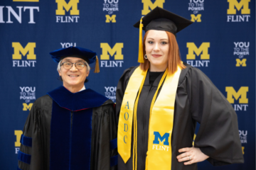 Two people standing in front of a UM-Flint banner background. Both are in graduation caps and gowns. On the left is Roy Barnes, AODC program director, and on the right is Alizia Hamilton.