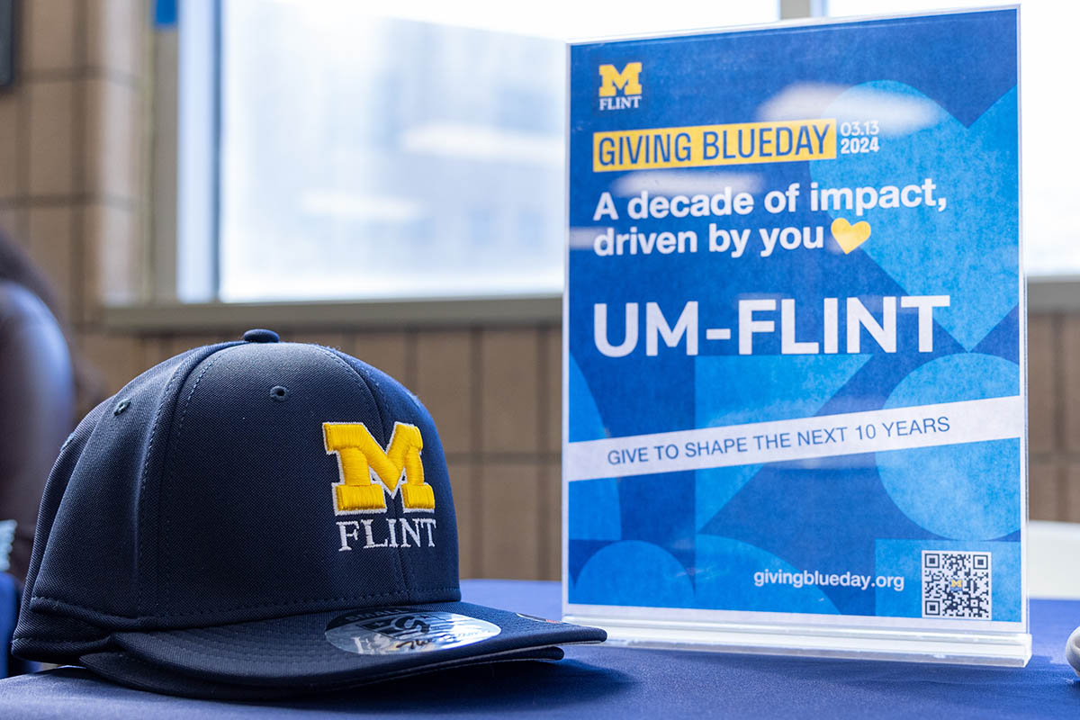 A UM-Flint baseball cap on a table with a Giving Blueday sign